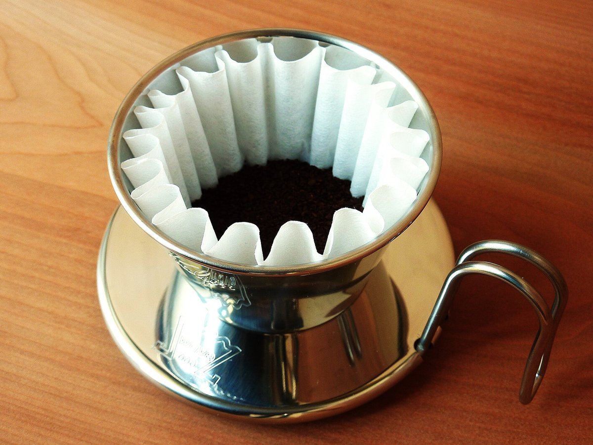 A stainless Kalita Wave dripper with its fluted paper filter and a bed of ground coffee, on a wooden table