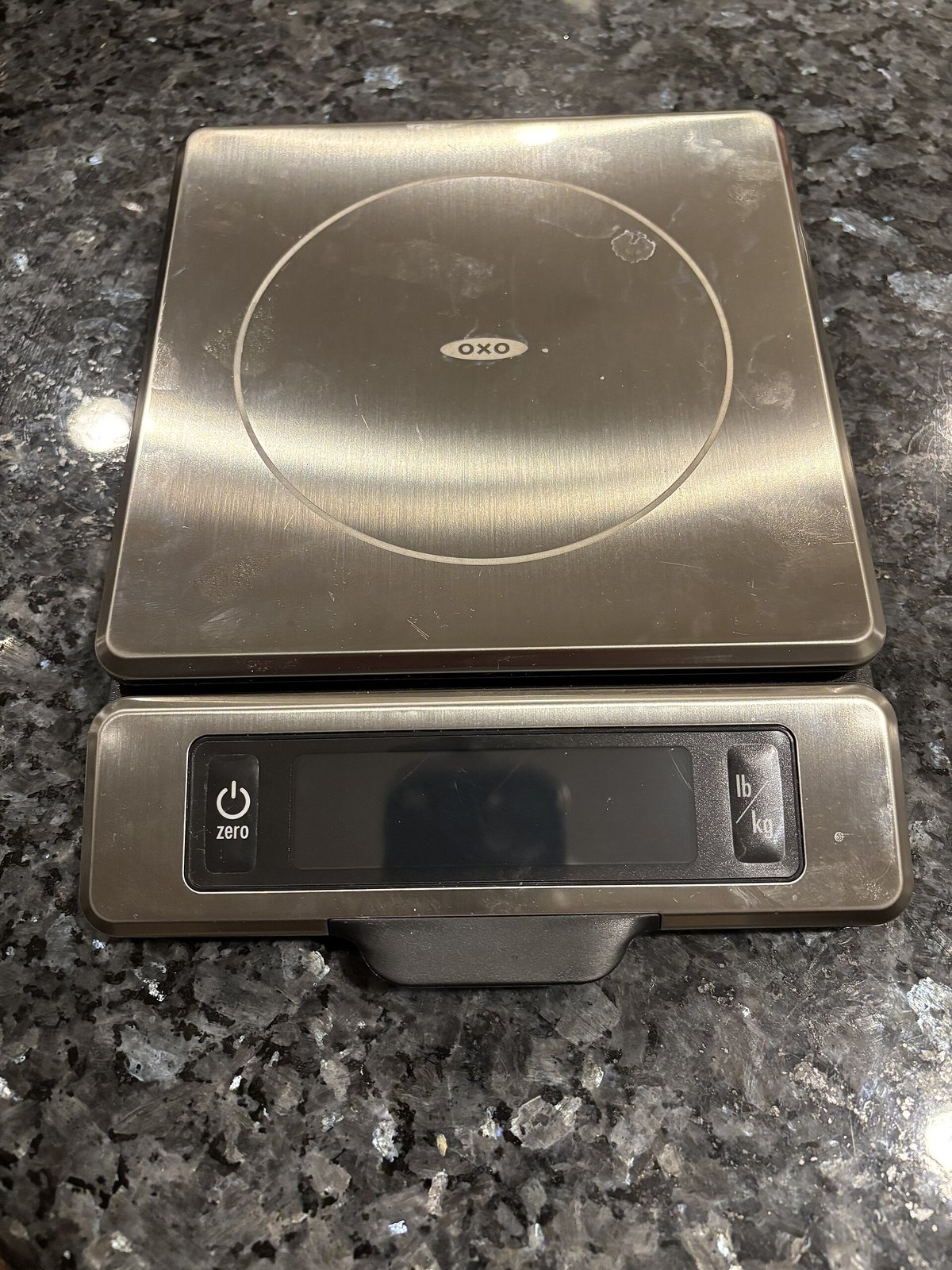 A square stainless kitchen scale with a grey display, on a granite counter