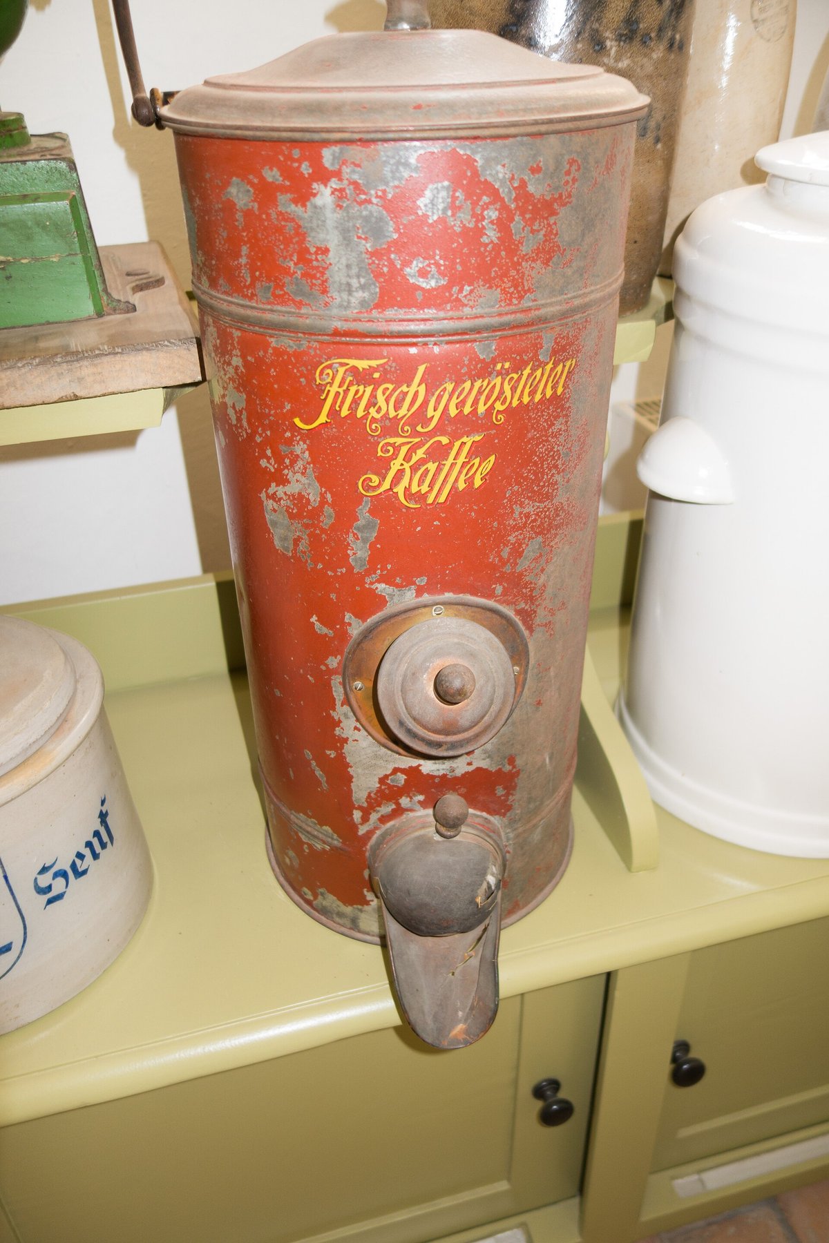 A tall red and cream tin coffee canister with a dispensing tap at the bottom, on a cream kitchen cabinet