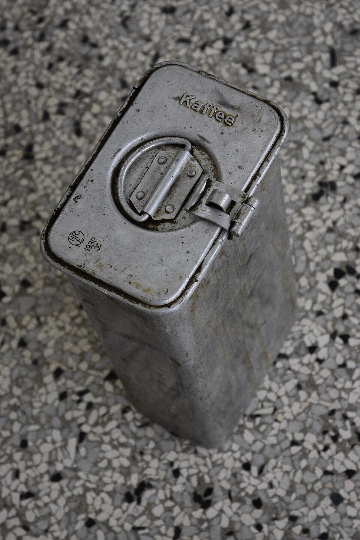 A battered aluminium coffee tin from 1938 with a clamp lid, standing on a speckled stone floor