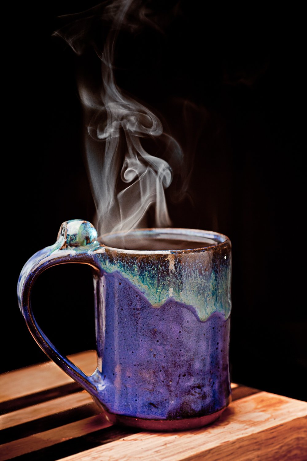 A steaming mug of black coffee in a glazed blue-green pottery mug on a wooden board, against a black background