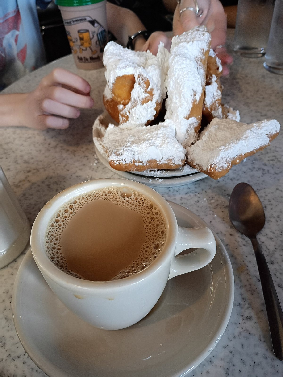 A cup of café au lait on a saucer beside a plate of beignets under powdered sugar on a marble table at Café du Monde, New Orleans, March 2026