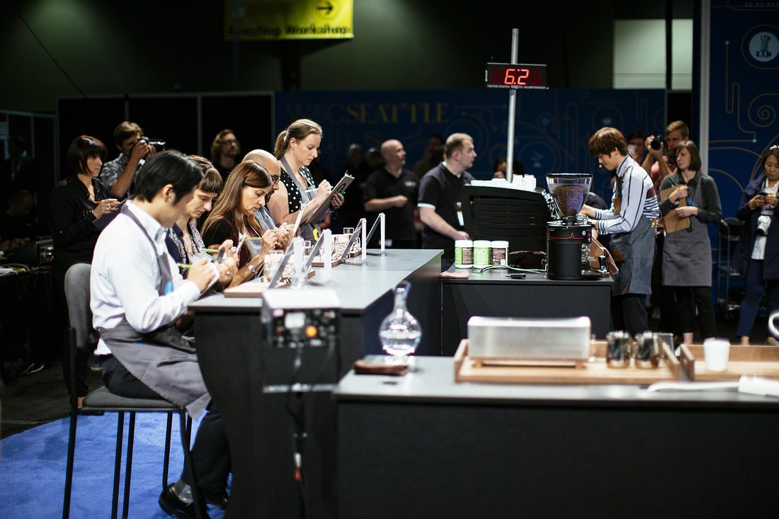 A competitor in an apron serves drinks to judges seated at a counter on a competition stage, a countdown clock behind