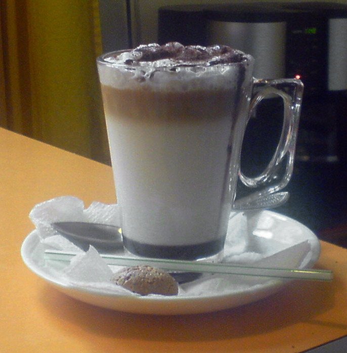 A tall glass mug of mocha in layers, milk below and a dark coffee band above, topped with froth and chocolate, on a saucer with a small cookie