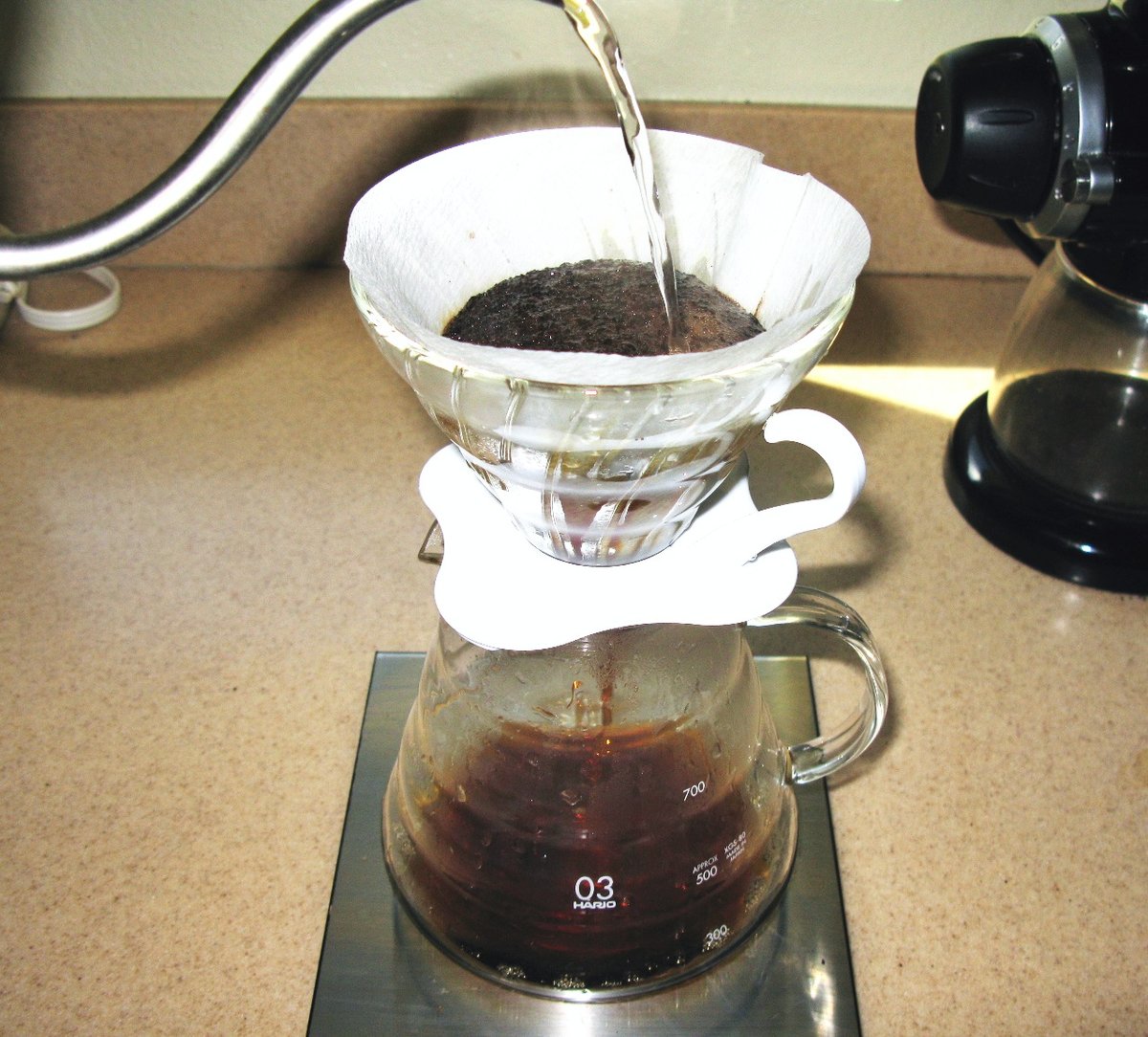 A glass pour-over dripper on a glass server standing on a digital scale, a gooseneck kettle pouring water onto the grounds