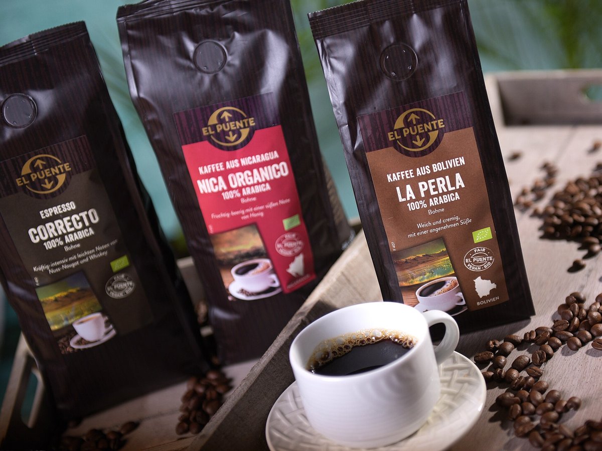 Three black coffee bags from the German fair-trade company El Puente, each with a one-way valve, the EU organic leaf logo and the company's own round fair-trade seal, beside a cup of black coffee