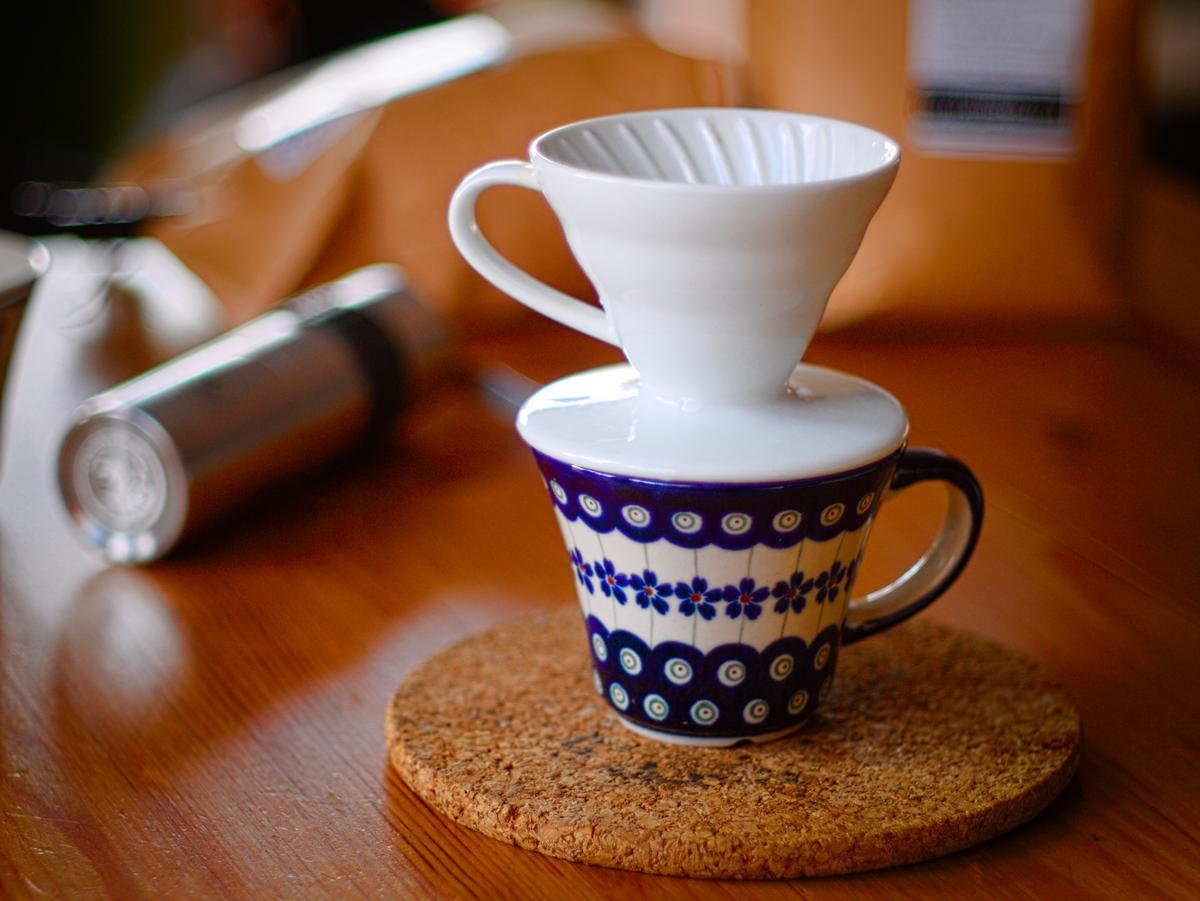 A white ceramic V60 cone dripper sitting on a blue and white patterned cup on a cork coaster, with a hand grinder lying behind it