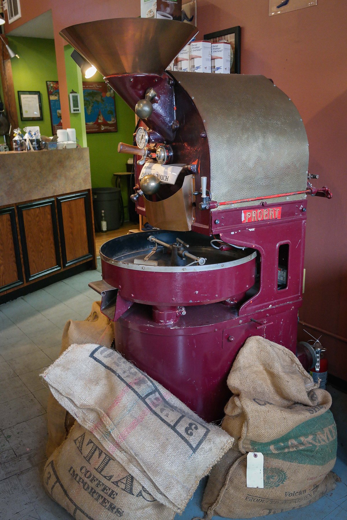 A shop roaster standing on its own sacks of green — hopper on top, drum in the middle, cooling tray out front. This is the machine behind most bags you buy from a local roaster