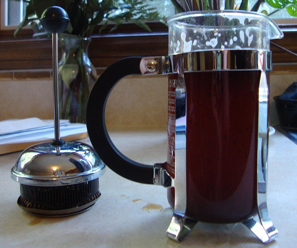 A glass French press full of dark brewed coffee on a table, its plunger and lid standing beside it