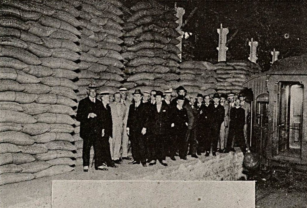 A black-and-white photograph of a group of men in suits standing in front of a wall of coffee sacks stacked to the ceiling