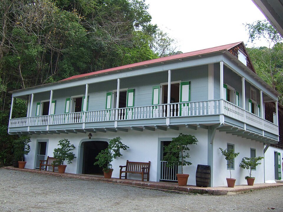 A two-storey plantation house with a long white balcony and a red tin roof, set against forest