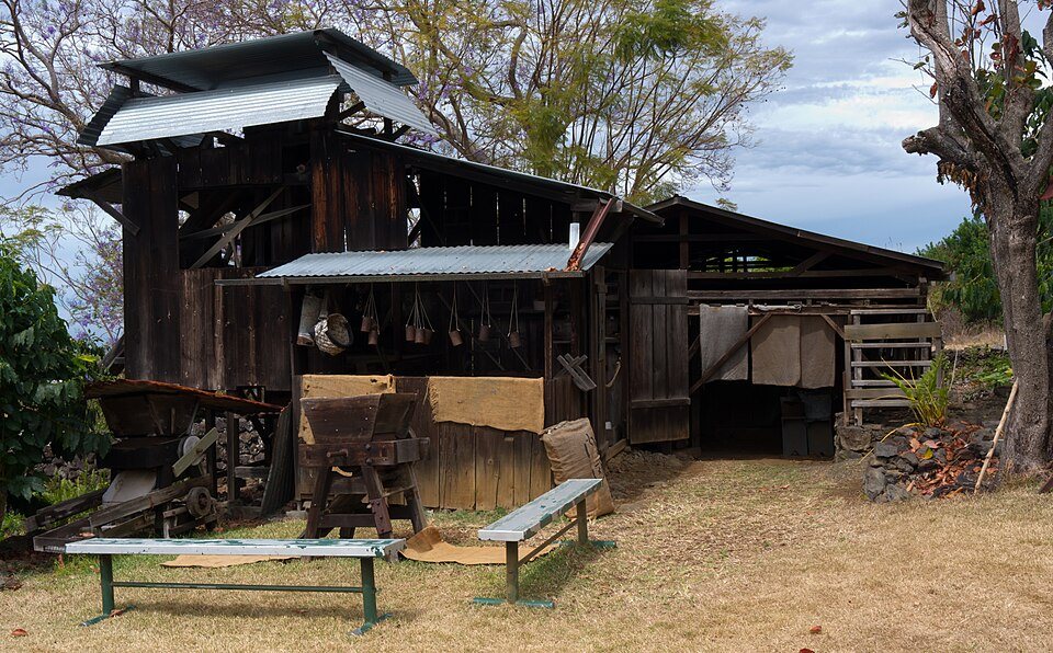 A weathered wooden mill building with a tin roof on a dry hillside, old machinery under its eaves