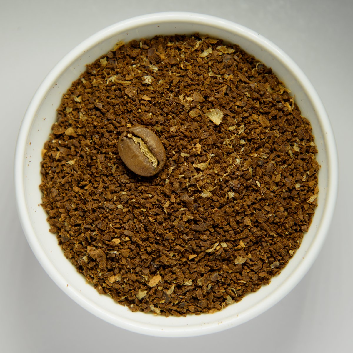 A white bowl of coarsely ground coffee with one whole roasted bean in the middle for scale