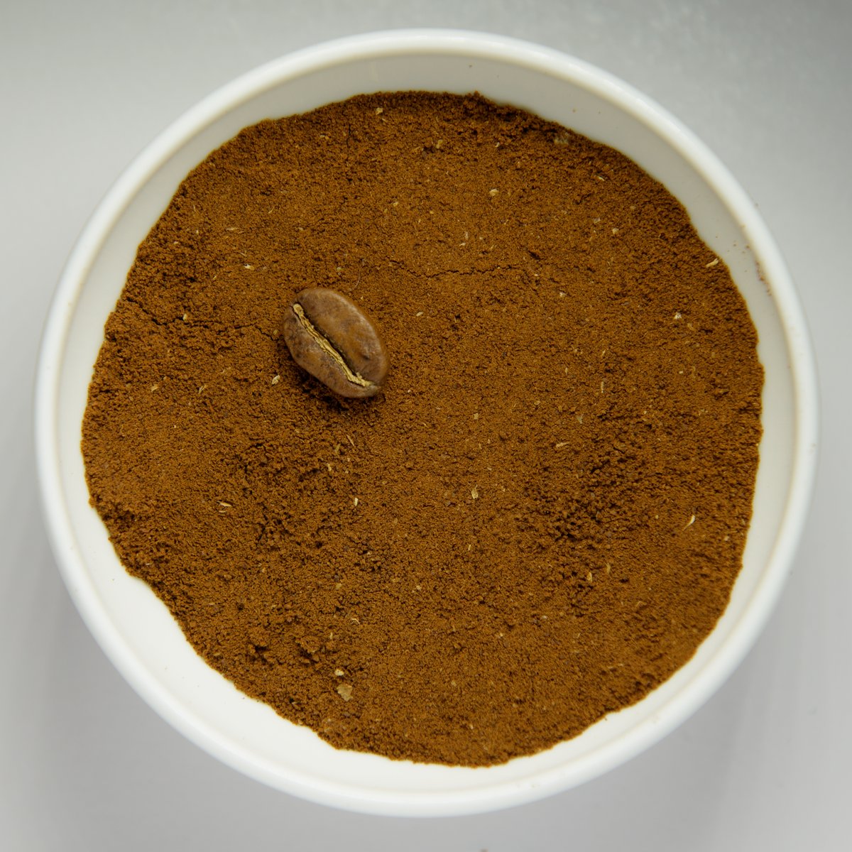 A white bowl of finely ground coffee with one whole roasted bean in the middle for scale