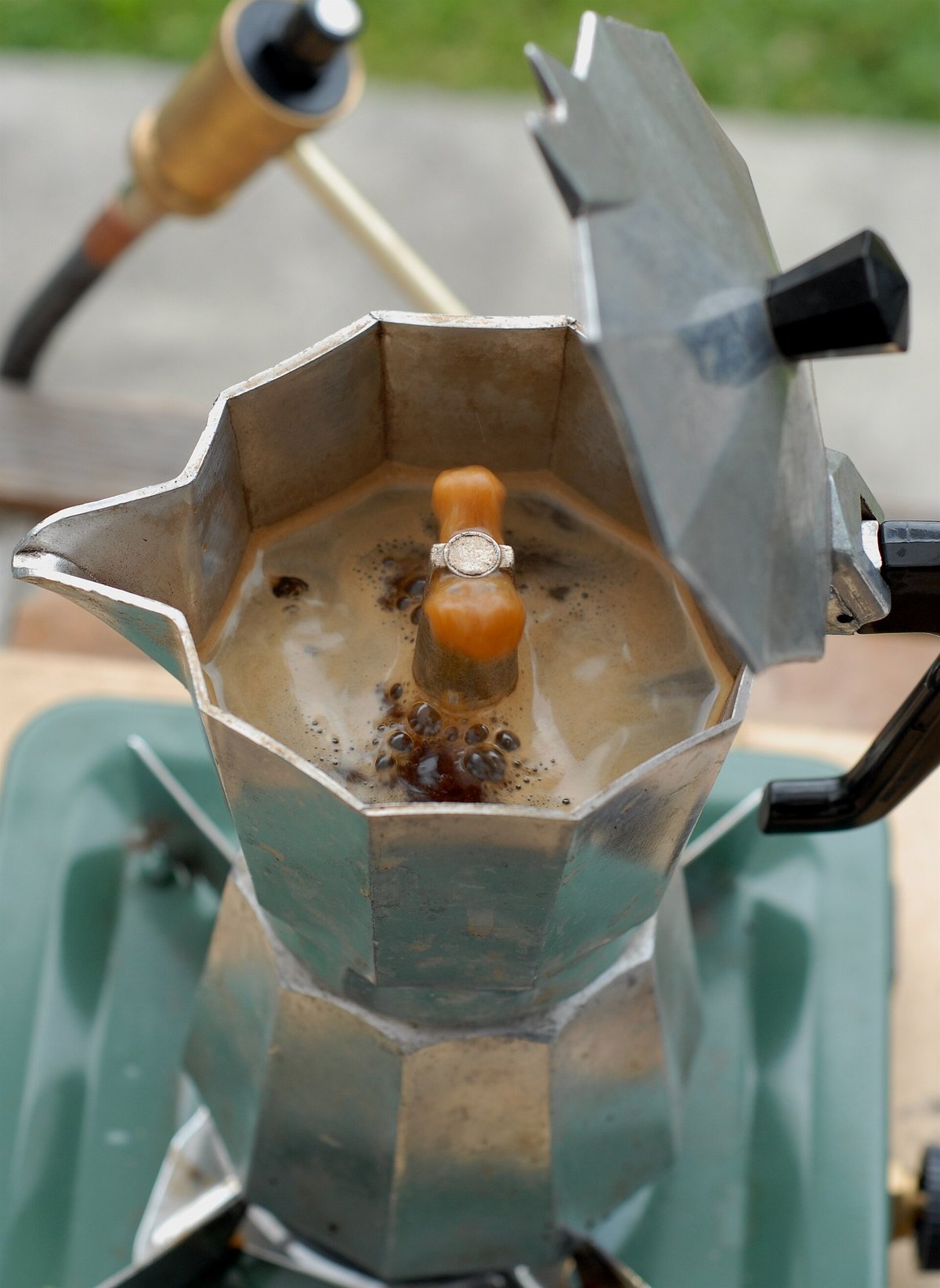 A stovetop moka pot on a gas burner with its lid open and coffee rising out of the central spout into the top chamber