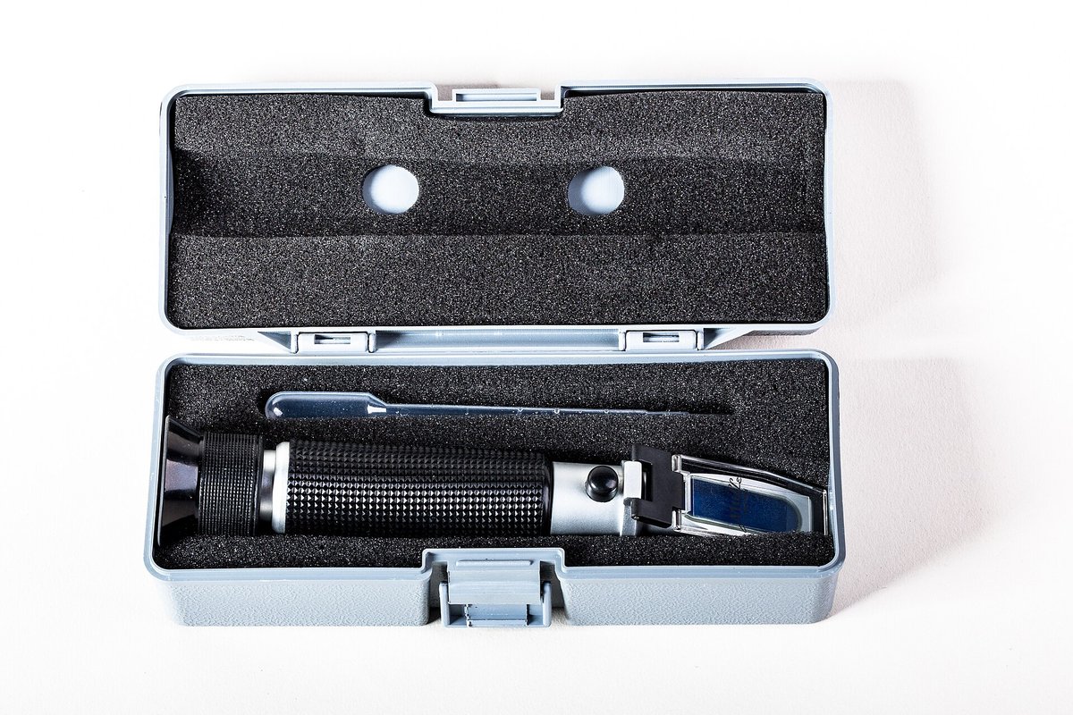 A handheld optical refractometer in an open grey carrying case with its black eyepiece and a small screwdriver
