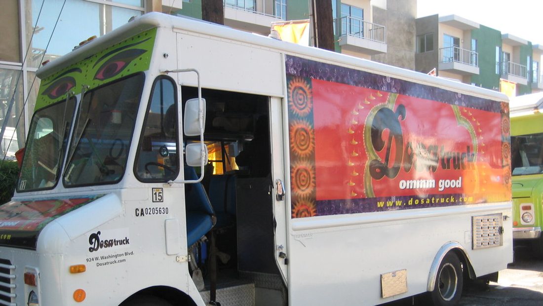 A food truck with its service window open, parked at a summer event with a small line of customers