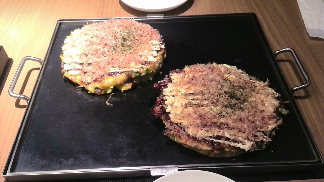 Two okonomiyaki finishing on a teppan, katsuobushi on top. Not captioned as any regional style — which is the argument of this page