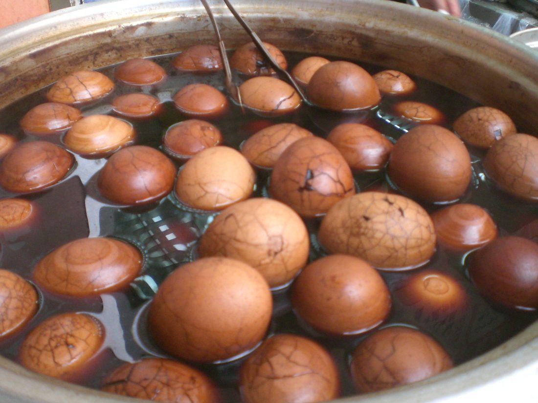 Tea eggs steeping in dark liquid in a metal pot, their shells cracked all over