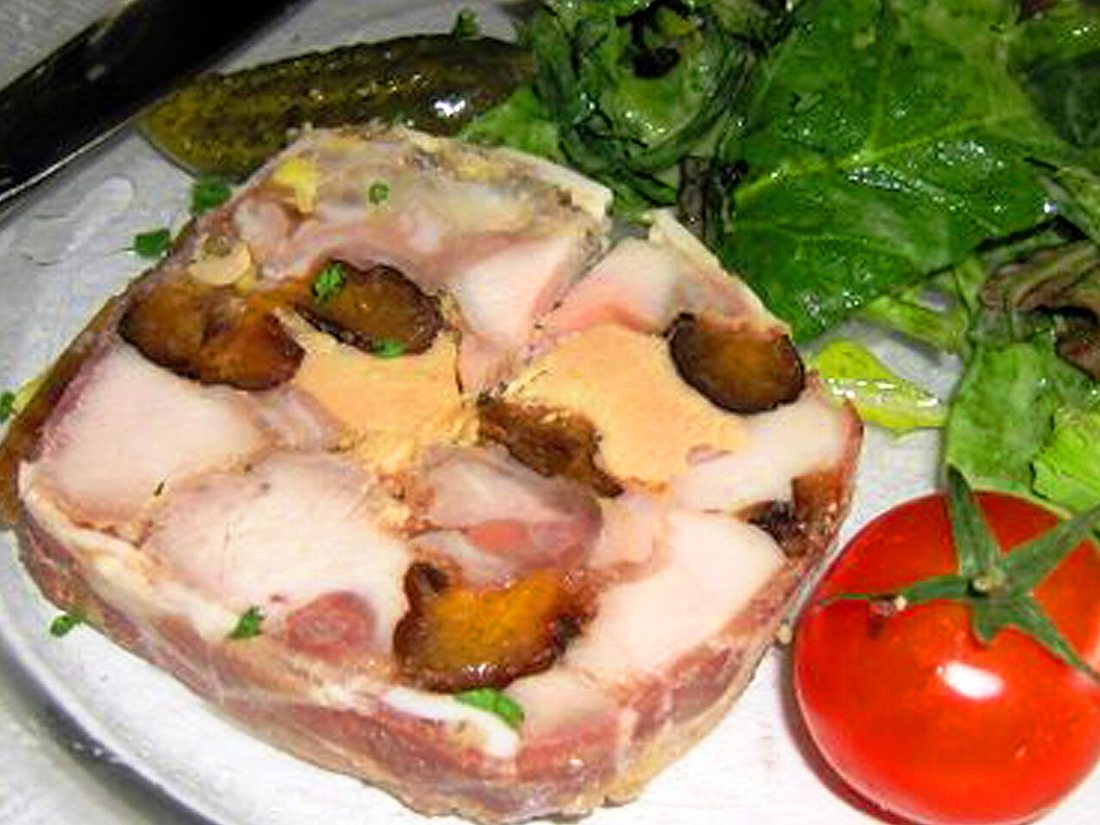 A slice of rabbit terrine showing large pieces of meat and dark fruit set in a lighter forcemeat, with a tomato and salad