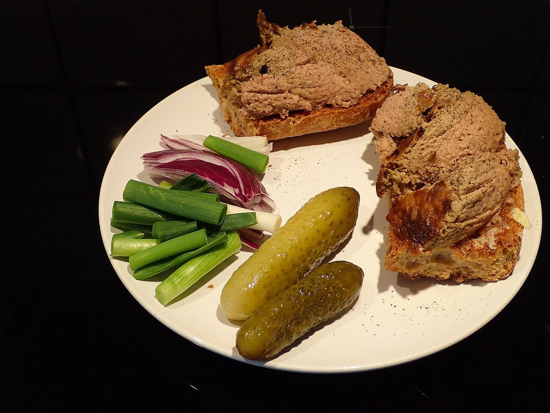Two slices of bread spread thickly with leverpostej on a white plate with pickles, spring onion and red onion