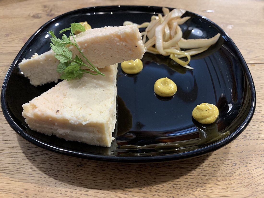 Two wedges of pale two-fish terrine on a black plate with dots of curry sauce and a garnish