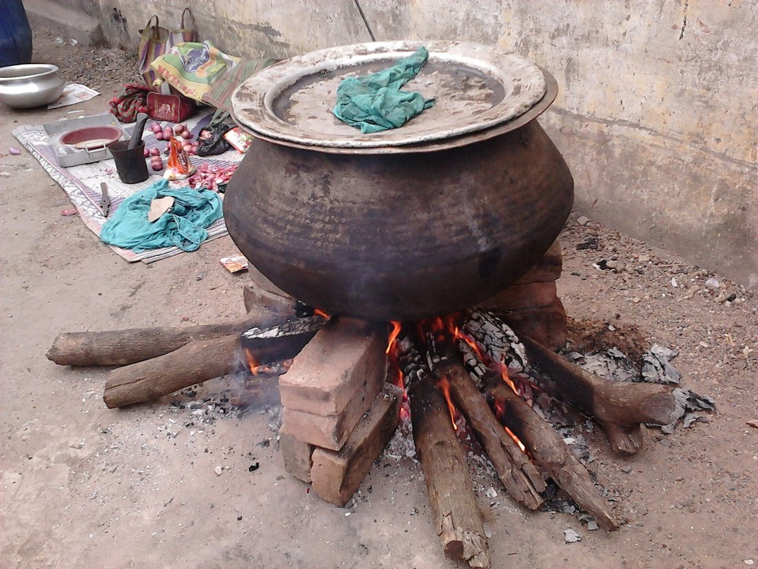 A large dark metal cooking pot on a brick-and-log wood fire in a street in Howrah, its wide lid weighted and covered with a cloth