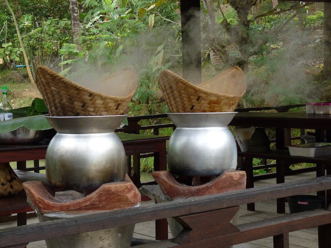 Two conical woven bamboo baskets set on aluminium pots over clay stoves, sticky rice steaming inside them
