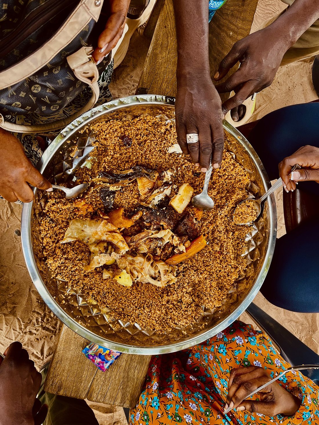 Seen from above, several people eating red thieboudienne with their hands and spoons from one large metal platter set on a mat