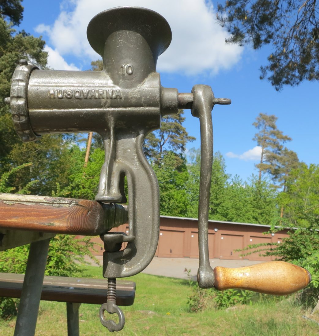 A 1902 Husqvarna No. 10 hand-crank meat grinder clamped to a table — the archetype of the cast-iron chopper that put a grinder in kitchens across Europe and America, hopper on top and the wooden-handled crank out to the side.