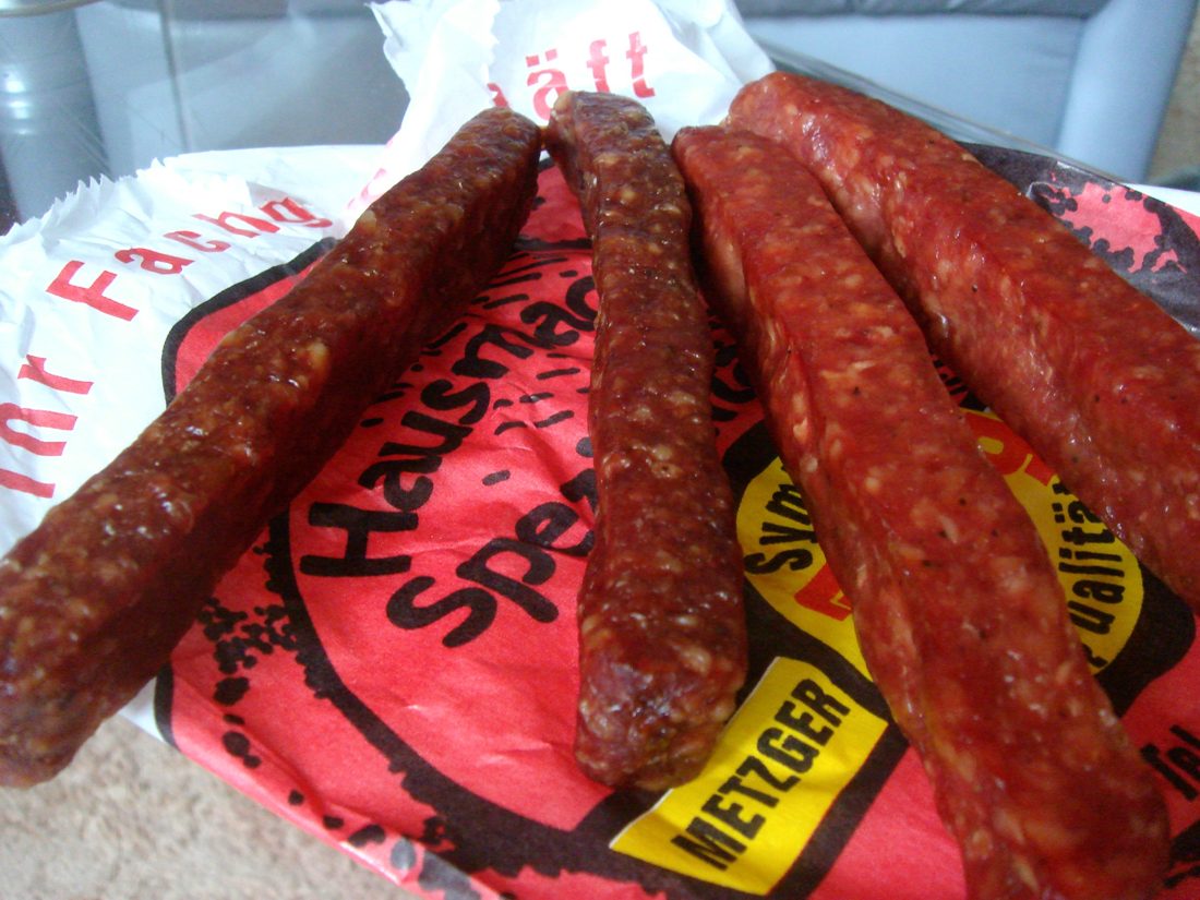 Close-up of Landjäger at a German butcher, a link cut across to show the coarse mosaic of ruddy meat and white fat inside a dark, tight-clinging casing — you can read the whole build from the cut face.