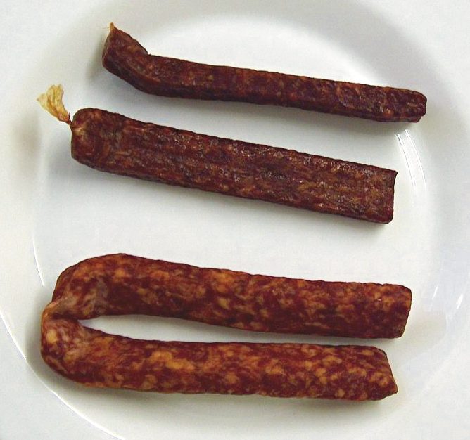 Three pressed pairs of German Landjäger on a white plate, one folded at the U-bend that joins the two links — this is the real thing, not a snack stick, and the flat brick profile is the whole point.
