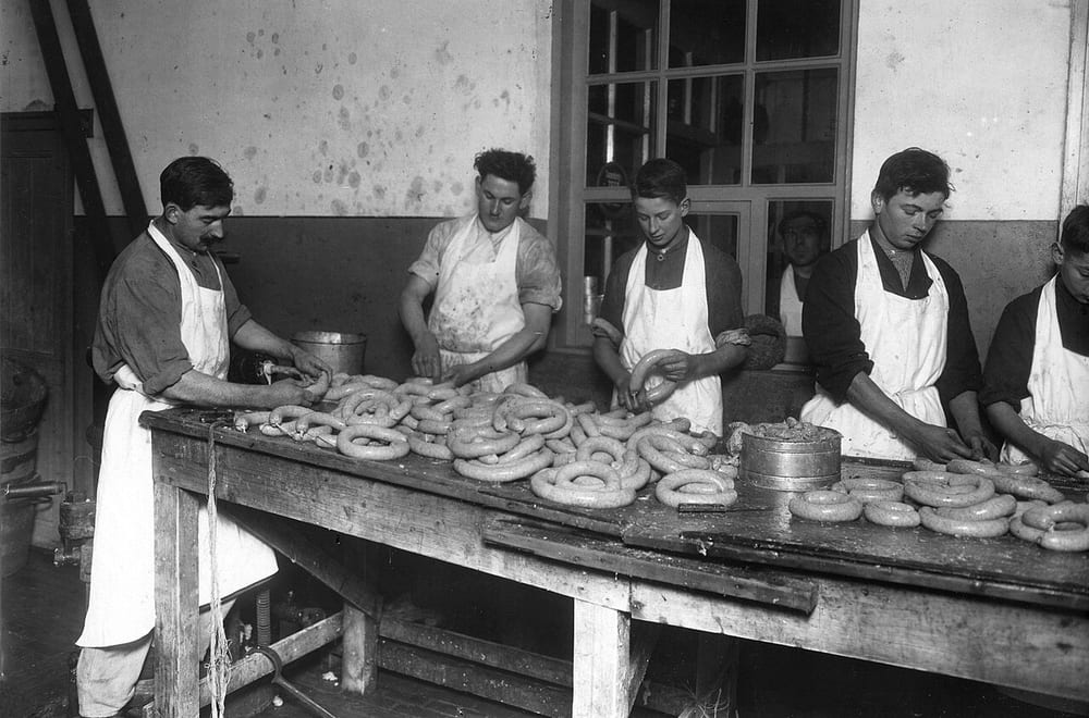 A communal pig butchery in progress, several people working at once around a long table — the blood-and-offal branch was never a one-person job, and the reason is a clock, not a custom
