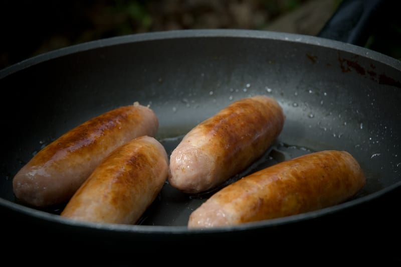 Pale plump bangers in a cast-iron pan, three of them just turning golden, visibly softer and rounder-shouldered than a coarse continental link