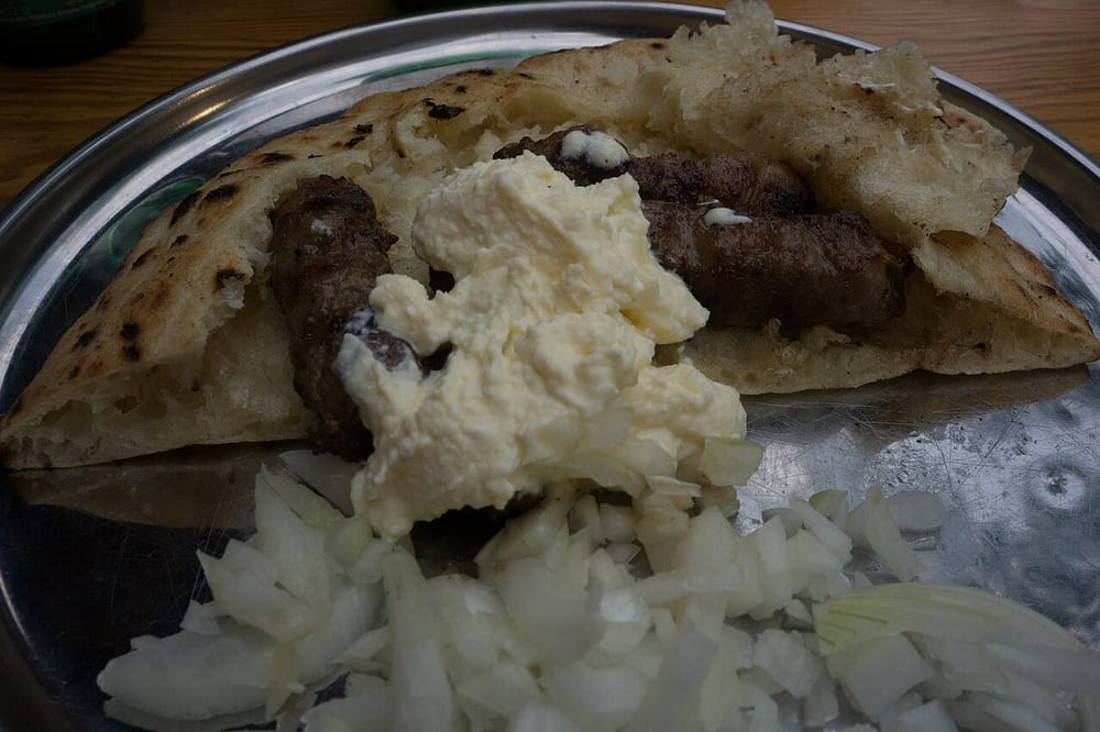 A dozen skinless ćevapi charred on a grill, packed into split flatbread with raw onion and a spoonful of kajmak