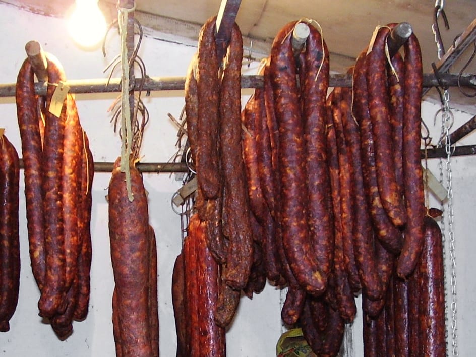 Two dark red Csabai sausages hanging in a smokehouse, dusted with paprika, indistinguishable by eye from a Spanish chorizo