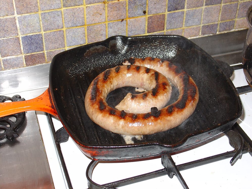 A yard of Cumberland sausage coiled flat in a black pan, unlinked, browned in a single continuous spiral with a skewer through it to hold the shape
