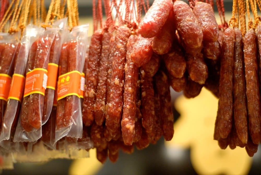 Pairs of lap cheong hanging on a rack, glossy dark red and visibly shrunken, the fat cubes translucent amber against the lean