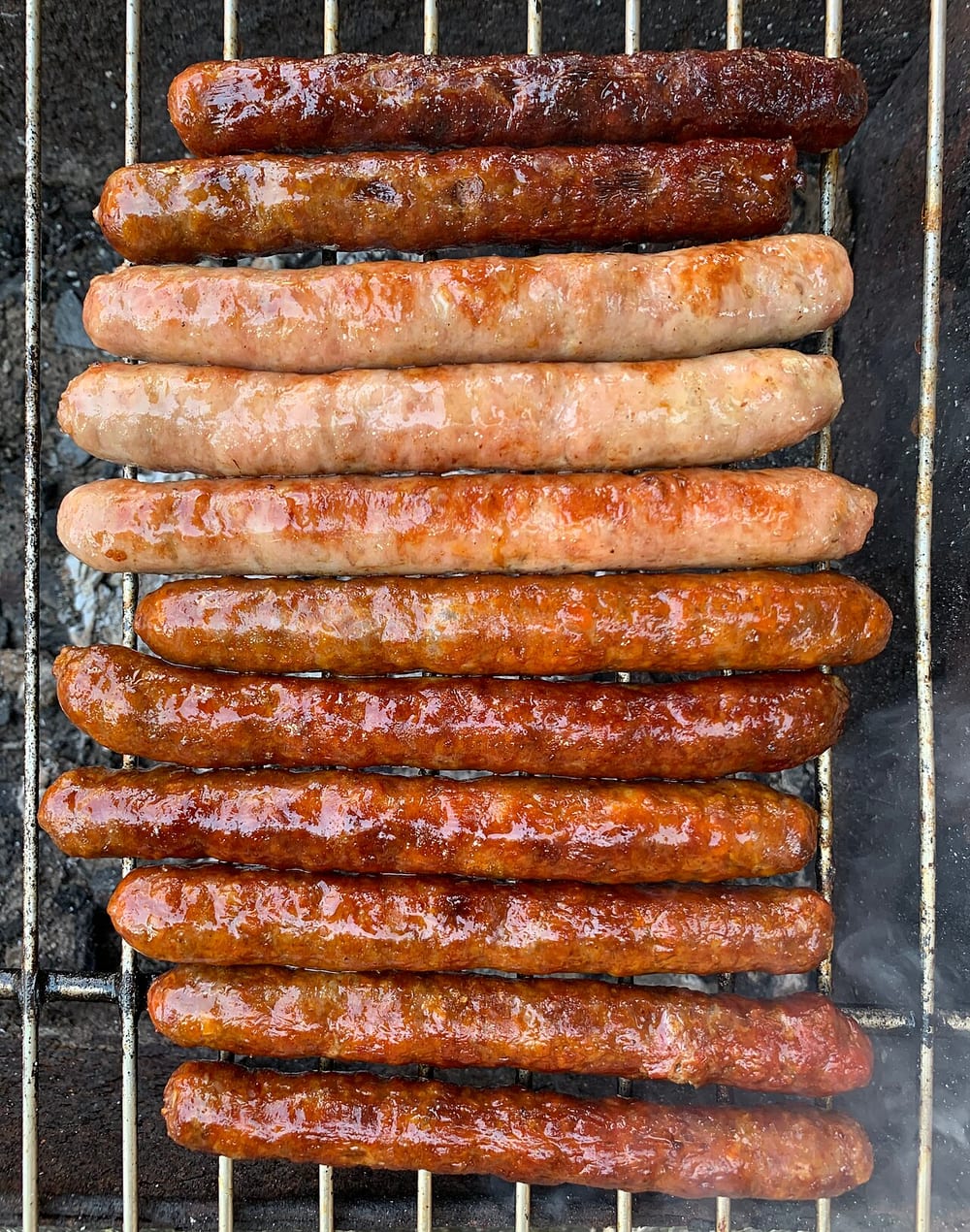 Thin dark-red merguez charring on a grill over the edge of the coals, blistered and glistening, noticeably slimmer than the links beside them