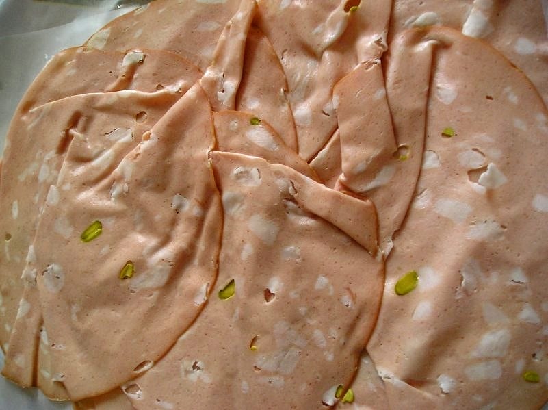 A single wide slice of mortadella draped over a hand, translucently thin, white fat cubes and green pistachios suspended in smooth pink