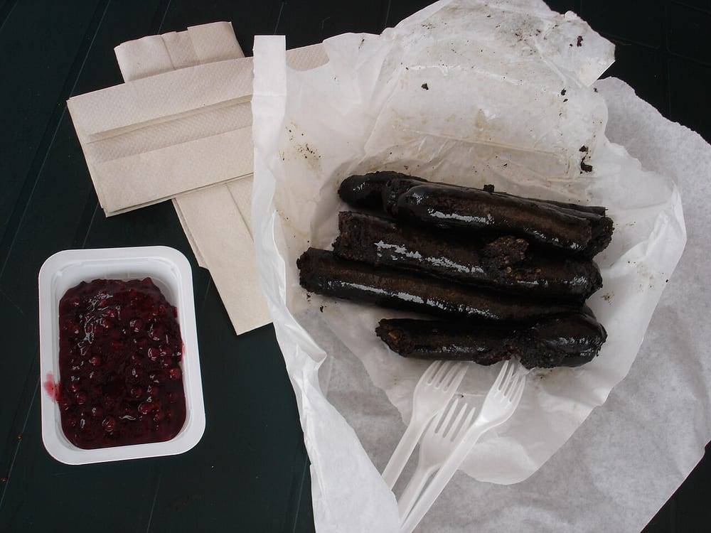 A coil of mustamakkara on a paper plate at a Finnish market stall, split open, a spoonful of bright red lingonberry jam beside it