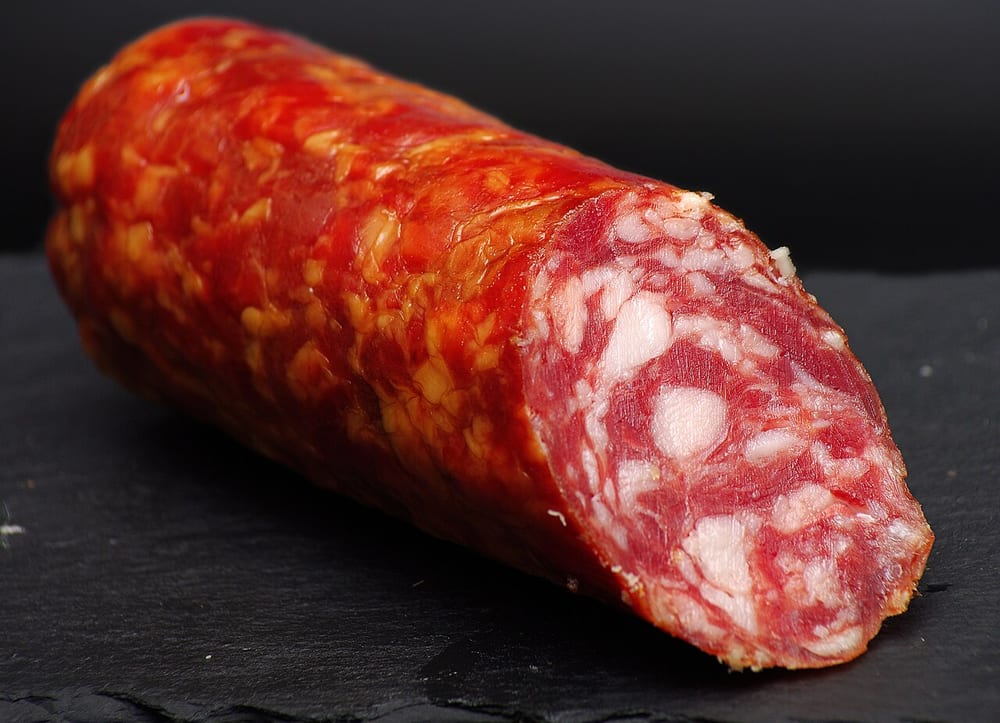 A slice of Salchichón de Vic held to the light, whole black peppercorns embedded through the pale pink paste like buckshot
