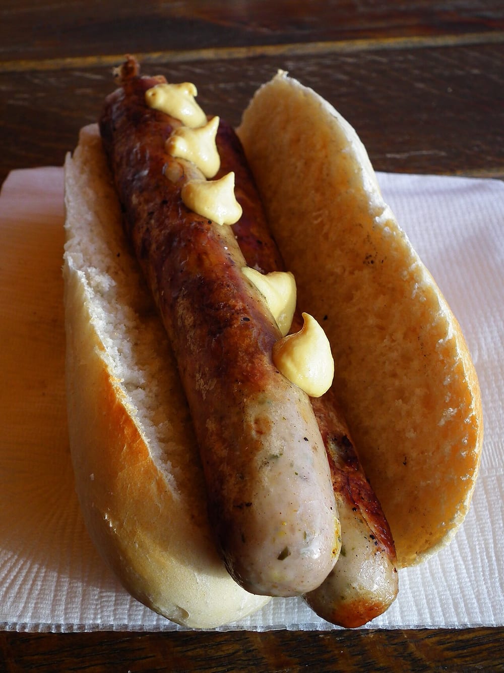 A long Thüringer bratwurst over glowing charcoal, easily 20 cm, the casing taut and unpricked, tucked into a split roll with mustard