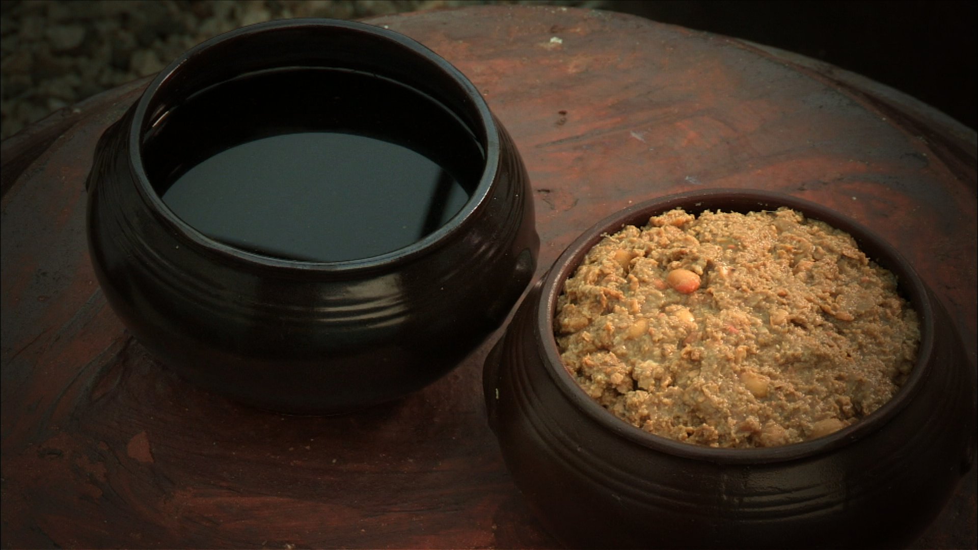 A dark crock of liquid ganjang standing beside a crock of thick brown doenjang paste