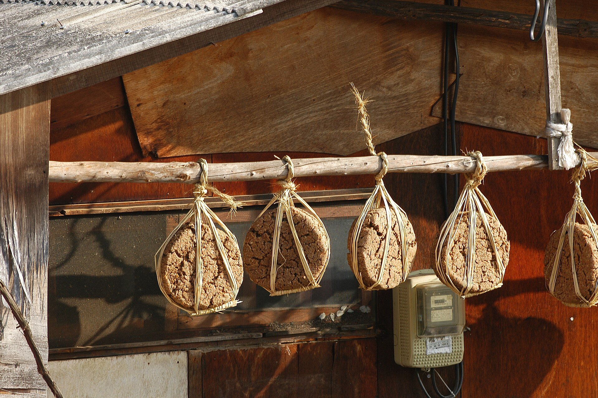Meju blocks bound in straw rope, hanging in a row from a wooden beam under a roof