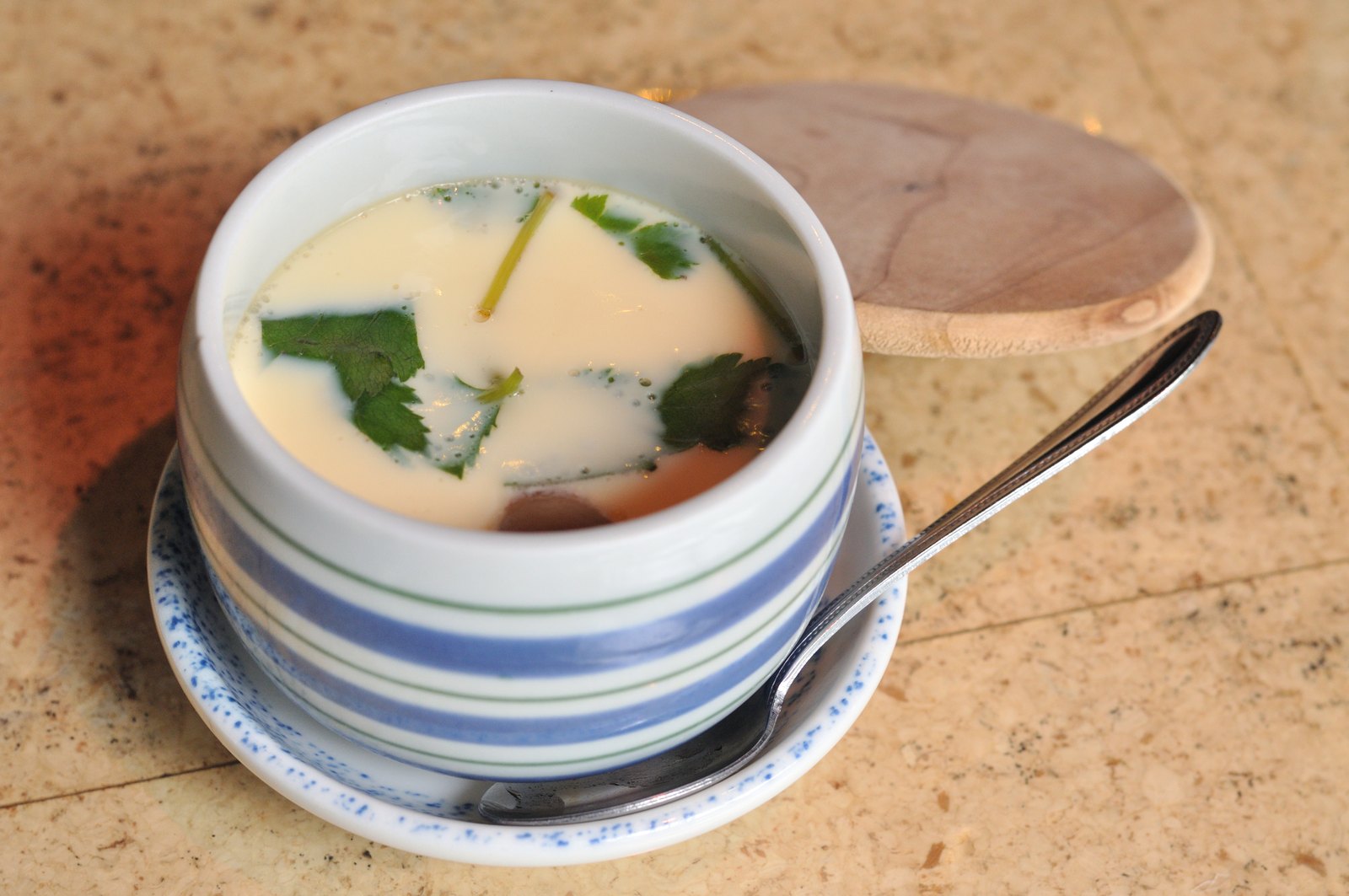 A chawanmushi cup with its lid set aside and the long-handled spoon on the saucer — the whole vessel, as it comes to the table