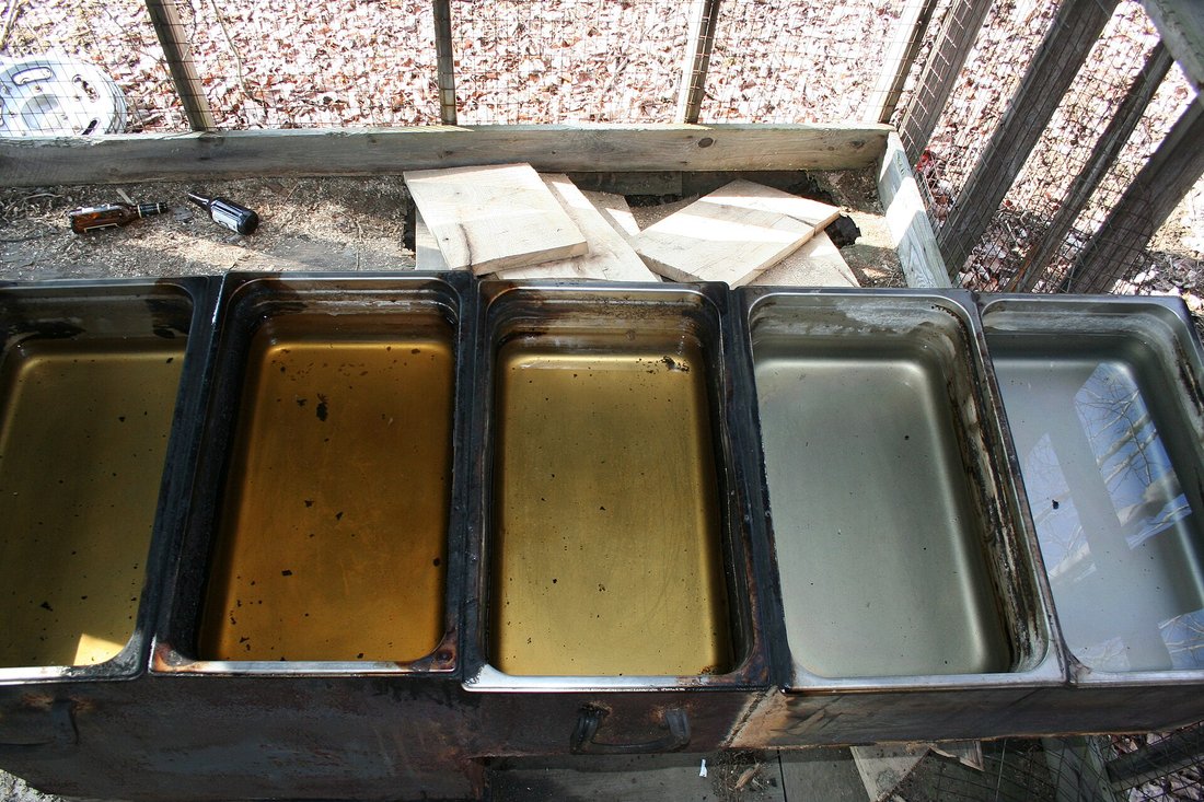 Looking down into a divided evaporator pan: five compartments side by side over the firebox, the sap in them running from nearly colorless at one end through pale straw to deep amber at the other — the gradient made visible