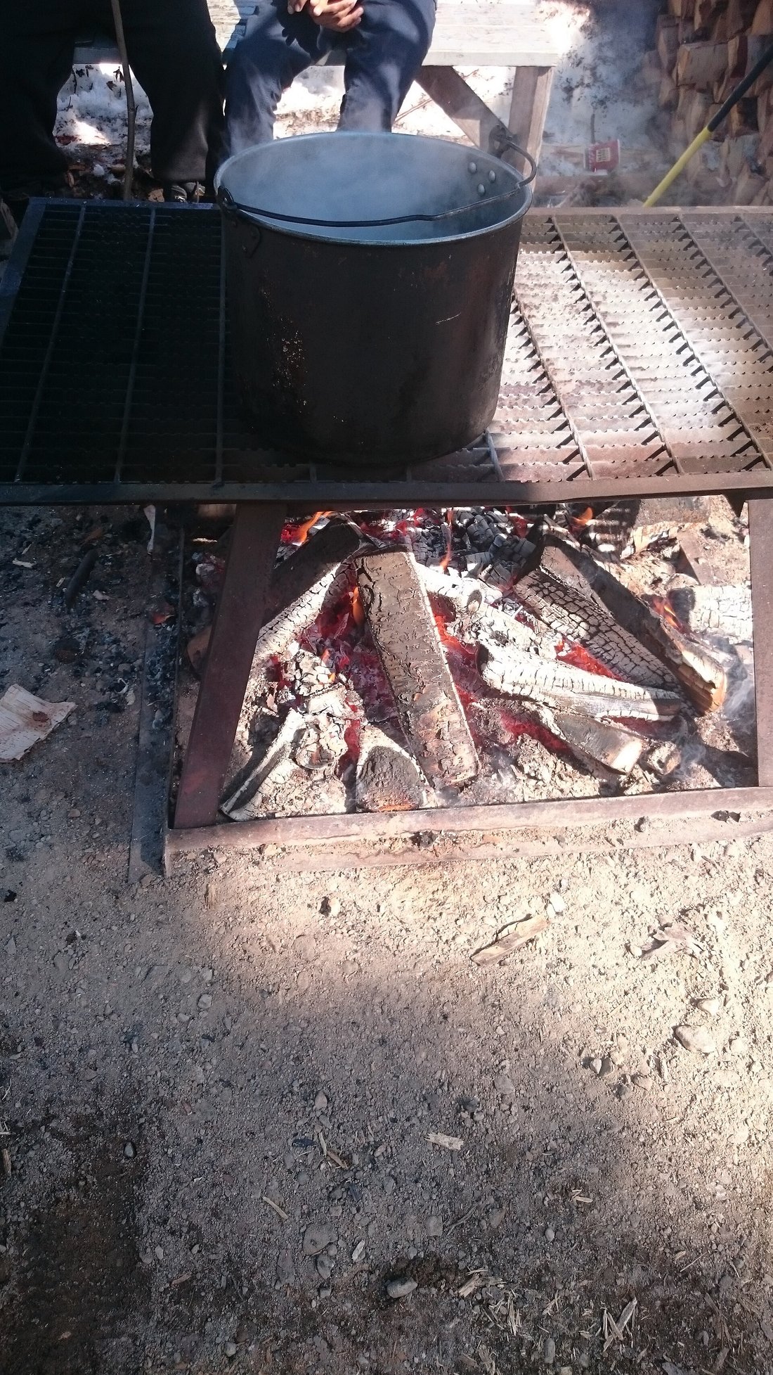 A pot of syrup coming up to temperature over an open wood fire, the fire pit cut into frozen ground with snow banked around it — the step before it goes onto the snow