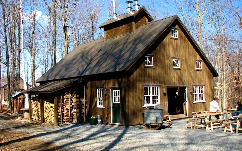 A working sugarhouse in winter: a small gable-roofed building with a long louvered cupola running the ridge, firewood stacked in rows under the eave, and a galvanized gathering tank sitting out front near the door