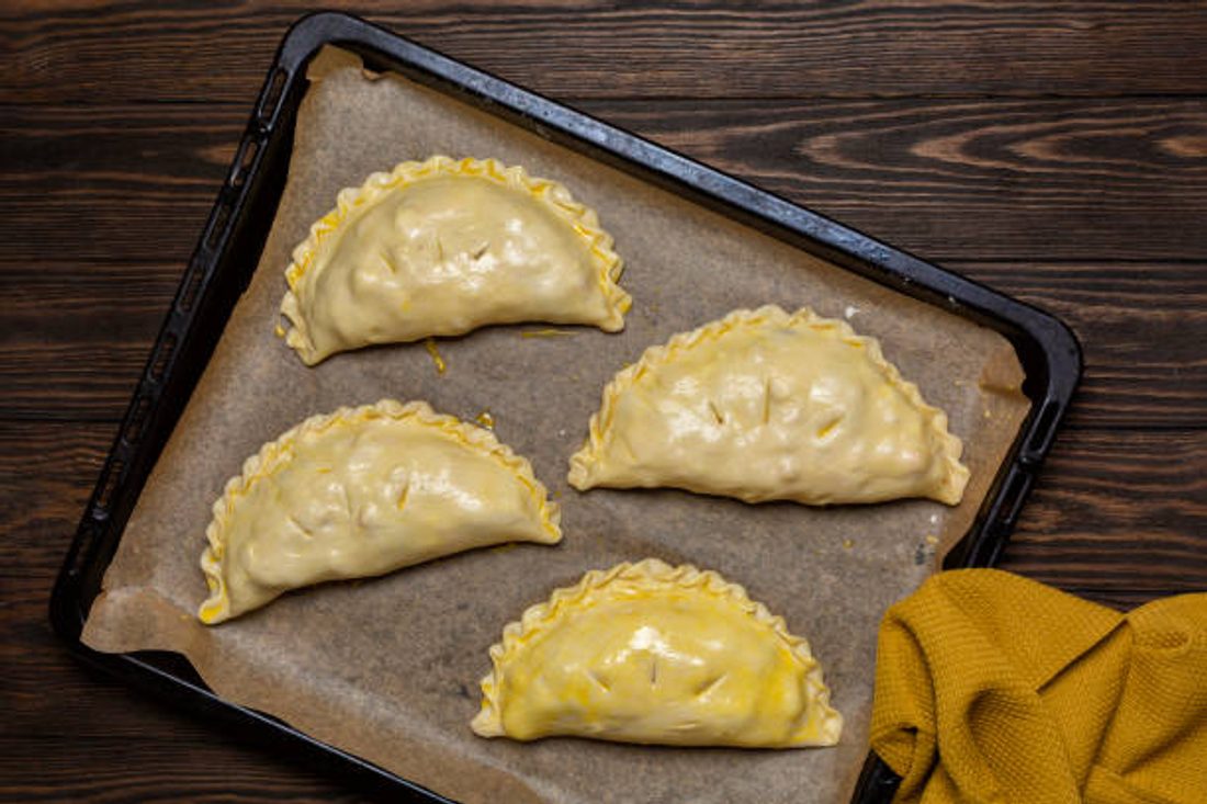 A tray of unbaked crimped pasties with egg wash and steam vents, ready for the oven